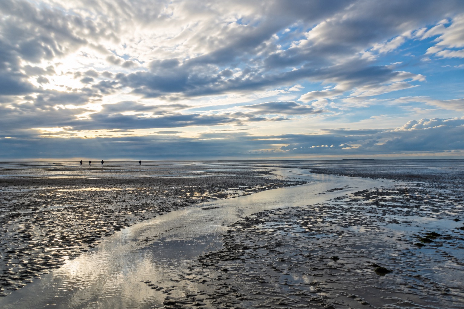 Wide exposed Wadden Sea mudflat at low tide under a dramatic sky, distant figures walking across the flats